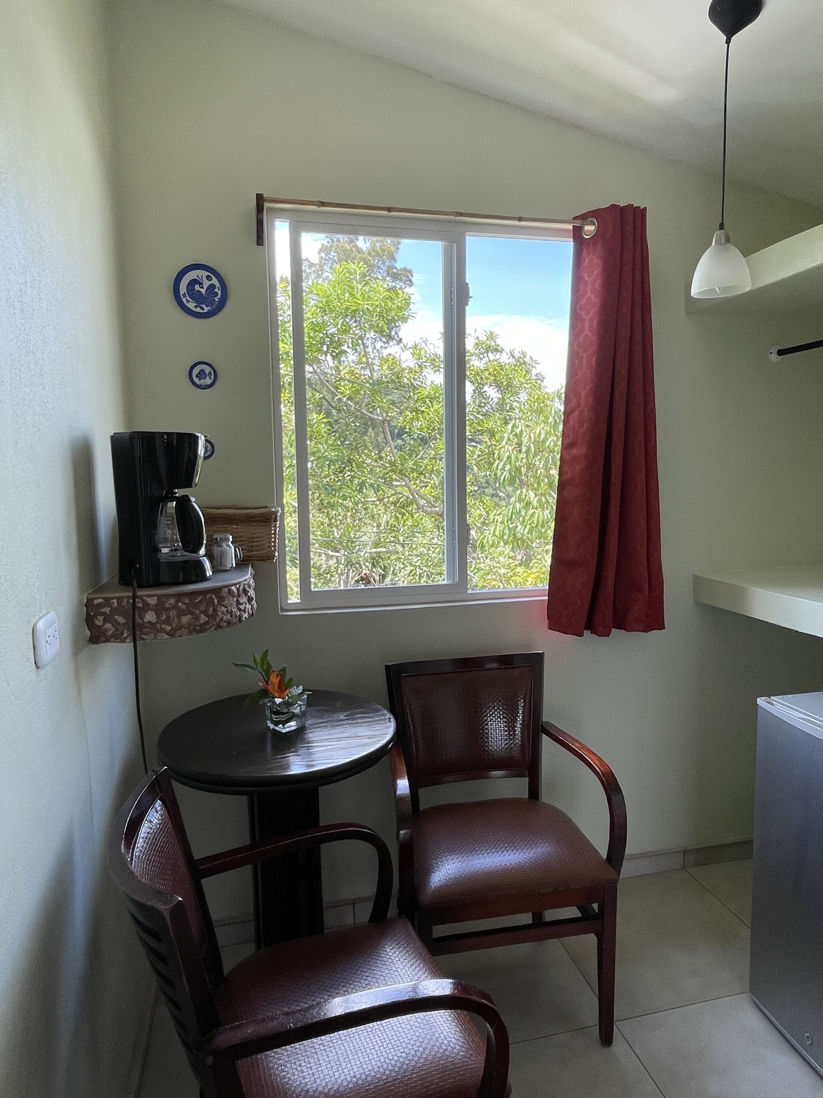 A small dining area with a round table and two chairs, a coffee maker on a shelf, a window with a red curtain, and a ceiling light.