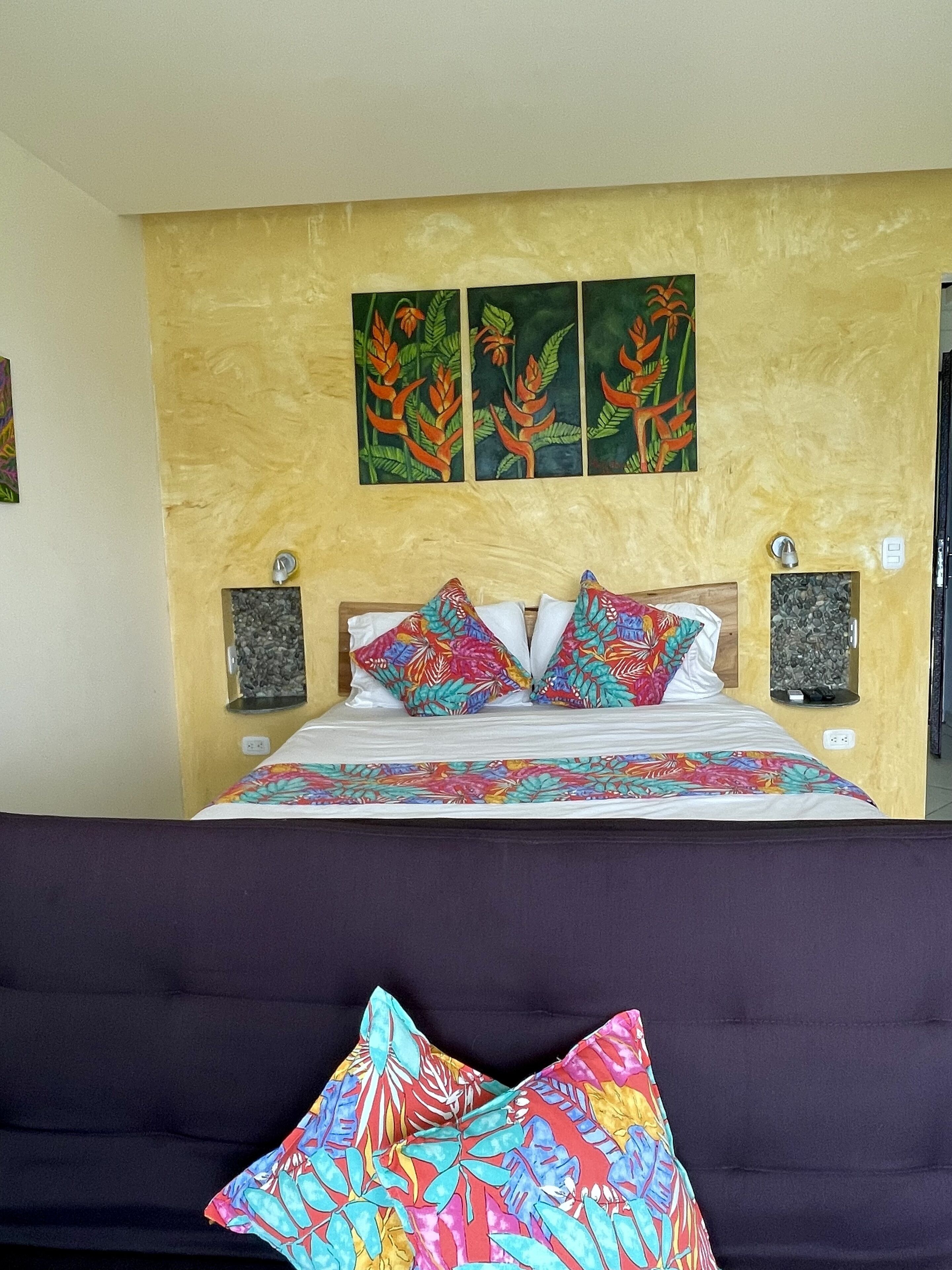 A bed with a floral bedspread, two pillows with a tropical pattern, and three wall-mounted paintings of tropical plants.