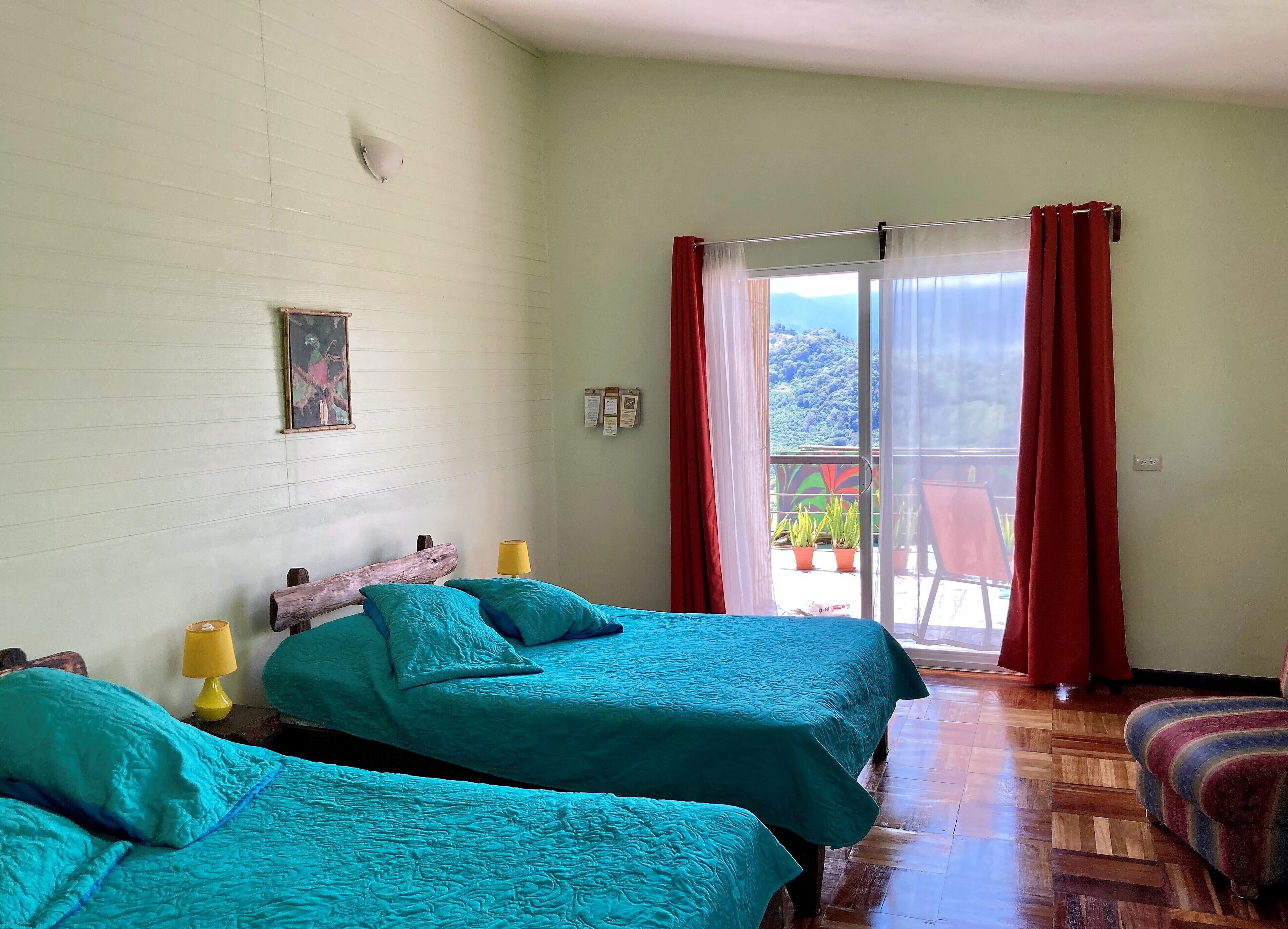 A room with two beds, a wooden headboard, a yellow lamp, a red curtain, and a view of a balcony with potted plants.