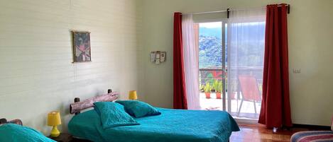 A room with two beds, a wooden headboard, a yellow lamp, a red curtain, and a view of a balcony with potted plants.