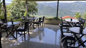 A covered outdoor seating area with tables and chairs, overlooking a mountainous landscape.
