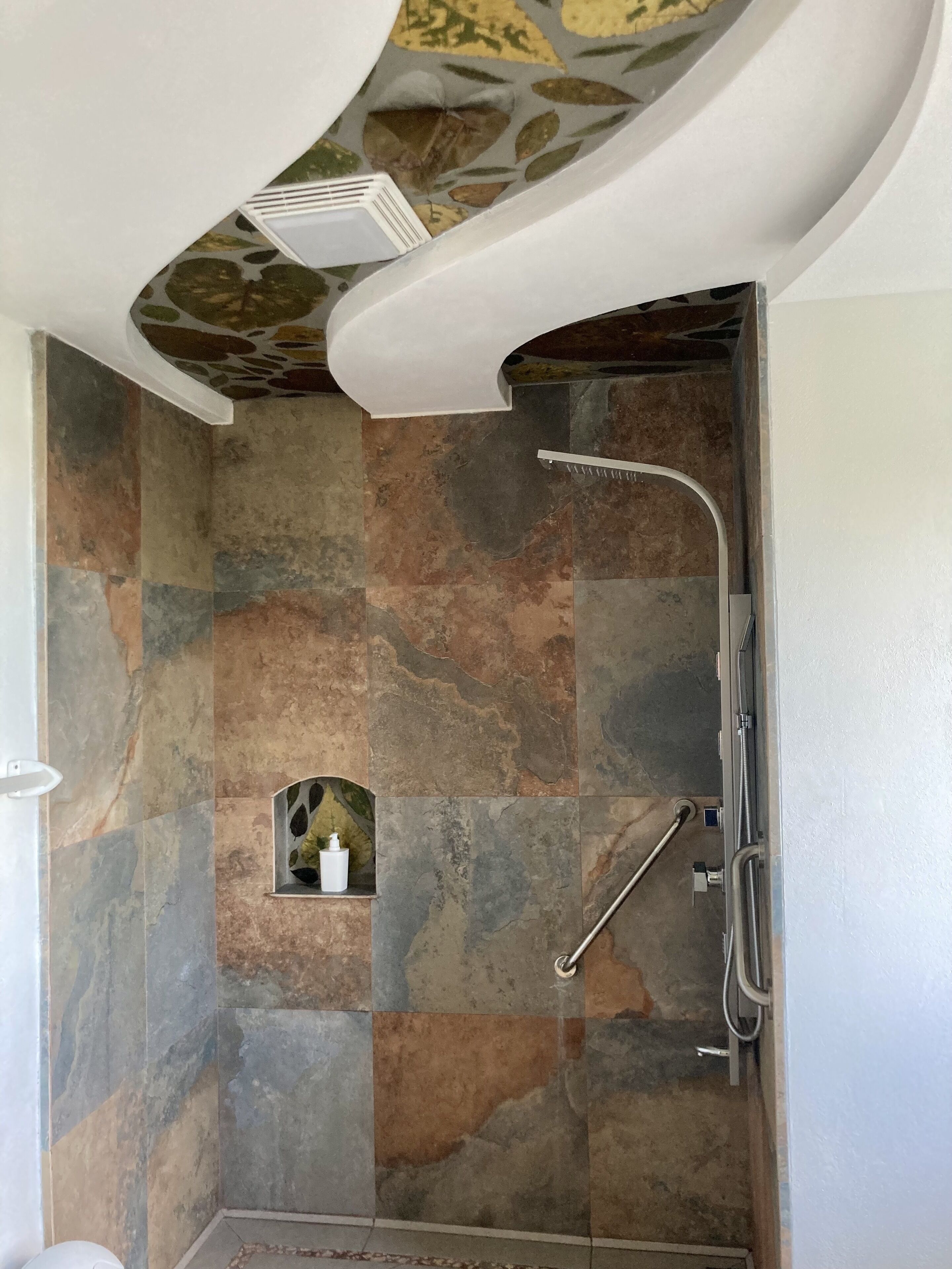 Ducha con techo curvo, azulejos de piedra y un estante pequeño con una botella.