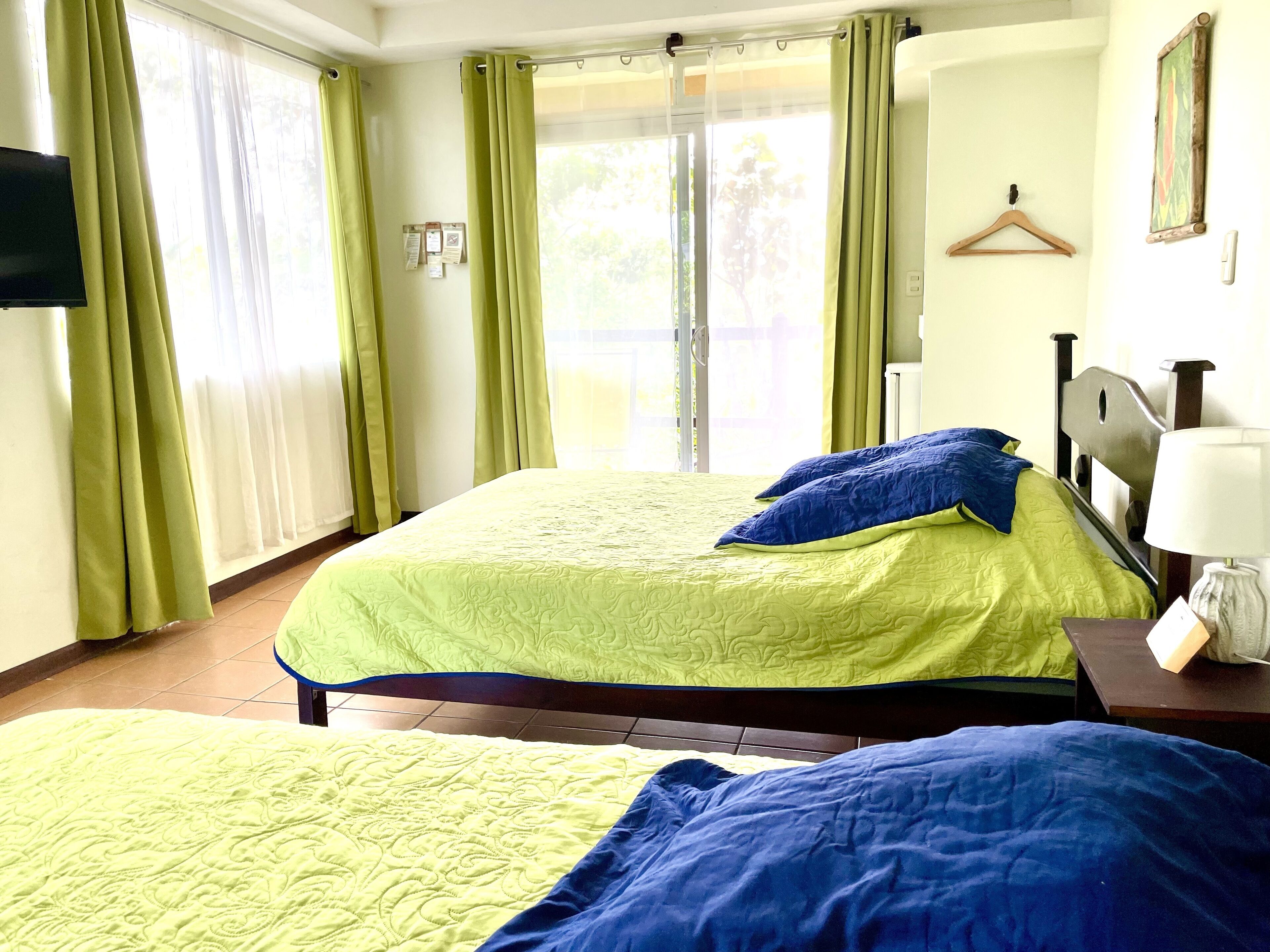 Habitación de hotel con dos camas, cada una con colcha verde y azul, cabecero de madera, mesita de noche con lámpara y ventana con cortinas verdes.