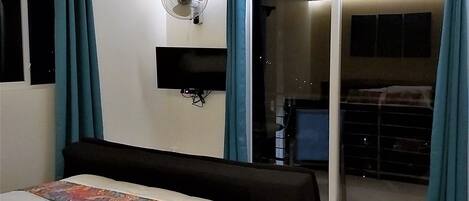A hotel room with a bed, a wall-mounted fan, a sliding glass door leading to a balcony, and a small TV on the wall.