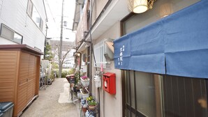A narrow alleyway with a building on the left and a storefront on the right. There are bicycles parked along the alley, a red mailbox, and a blue awning with Japanese characters.