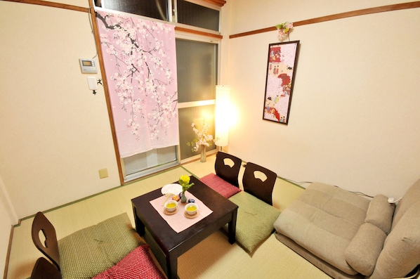 A traditional Japanese room with tatami flooring, a low table with a tea set, a sofa, and a window with a cherry blossom curtain.