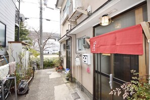 A narrow alleyway with a building on the right, featuring a red awning and a door with a handle. There are potted plants and a bicycle on the left.