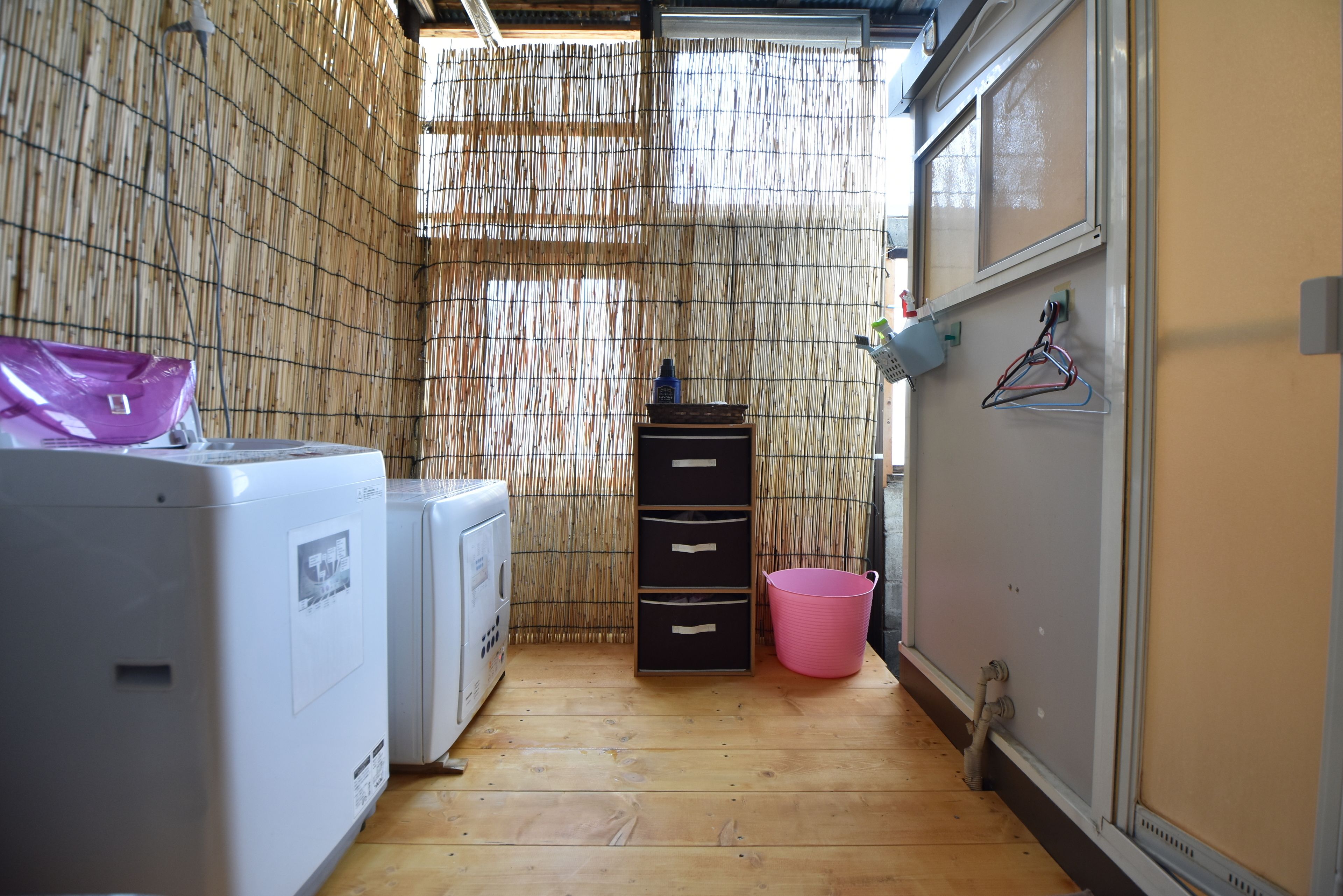 Una sala de bugaderia amb una rentadora, una assecadora, un sòl de fusta, un mur de bambú, un cubet rosa i una prestatgeria penjada a la paret amb penjadors.