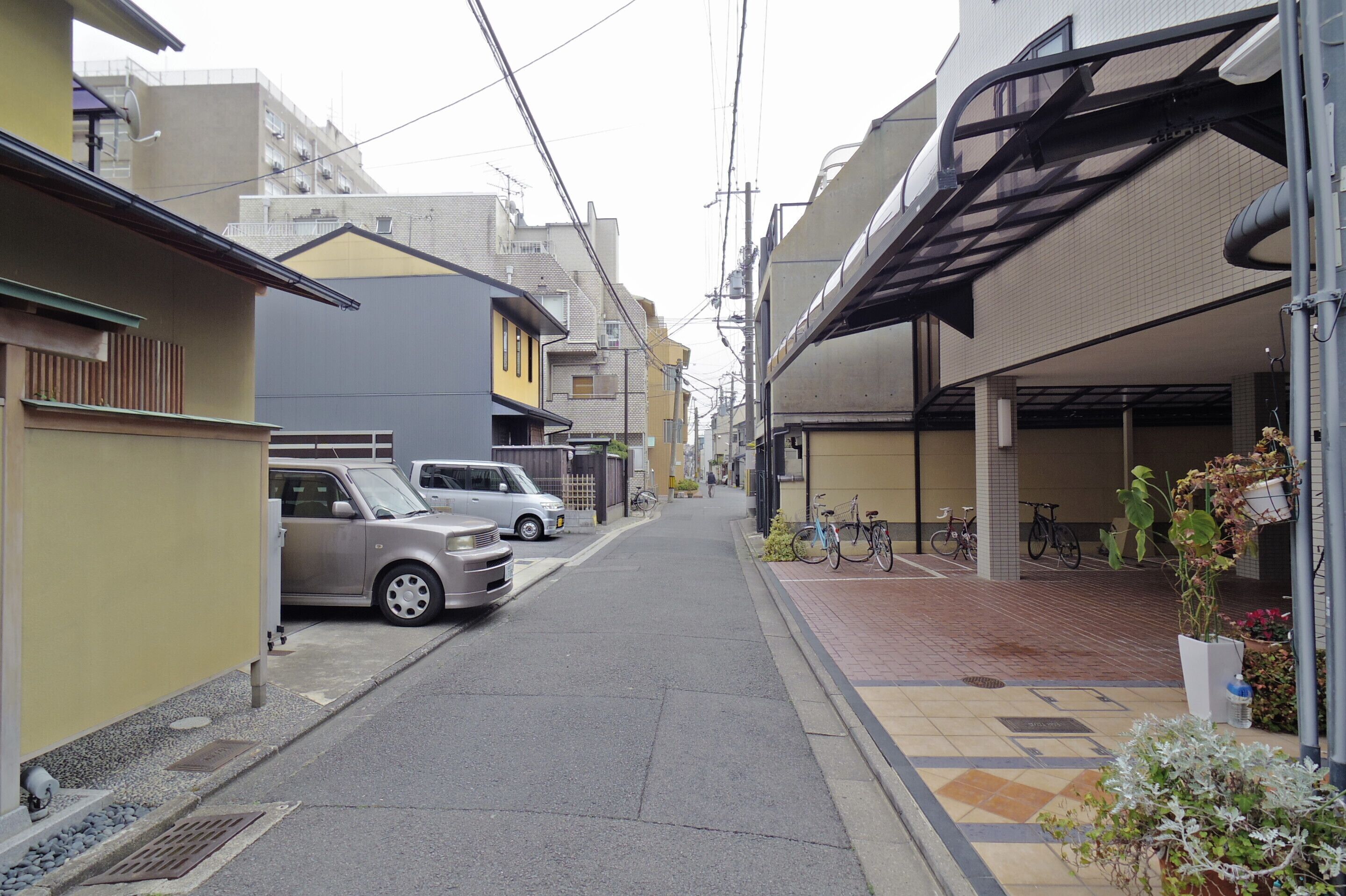A narrow urban street with parked cars and bicycles, buildings with balconies, and a pedestrian walkway.