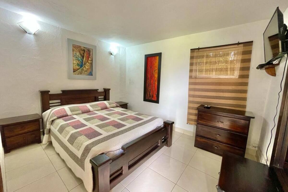 Une chambre à coucher comprenant un lit, une table de chevet, une commode et une fenêtre avec des stores.