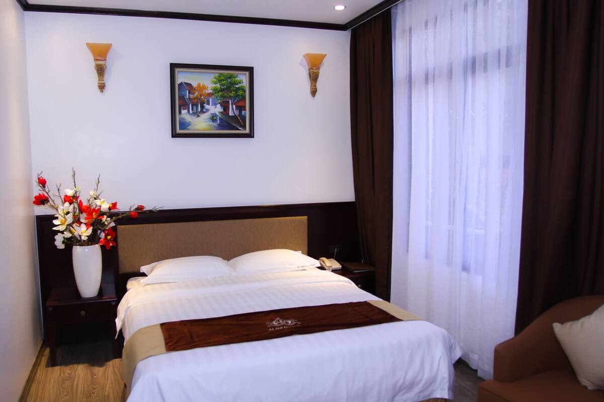 A hotel room with a large bed, a nightstand with a vase of flowers, a painting on the wall, and a brown sofa.