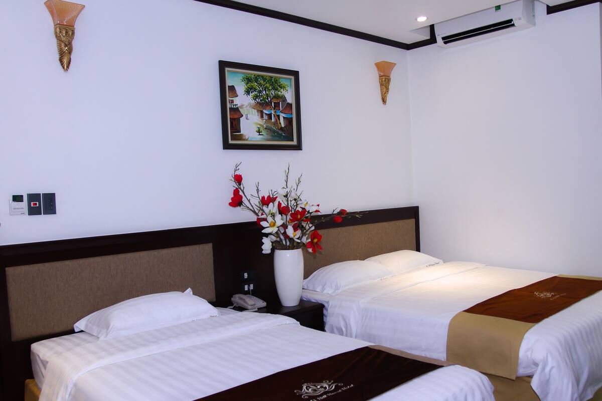 A hotel room with two beds, a nightstand with a phone, a vase with flowers, and a painting on the wall.