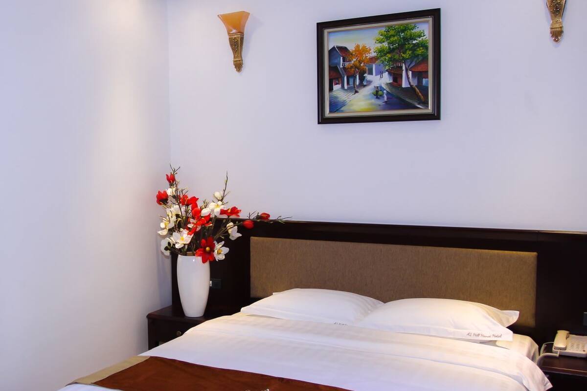 A hotel room with a bed, a vase of flowers, a painting on the wall, and a nightstand with a phone.