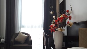 Quarto de hotel com uma janela grande, uma cadeira com almofada e um vaso com flores.