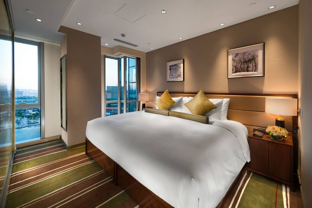 Ein Hotelzimmer mit einem großen Bett, einem Nachttisch mit Lampe und Blick auf die Stadt durch ein großes Fenster.