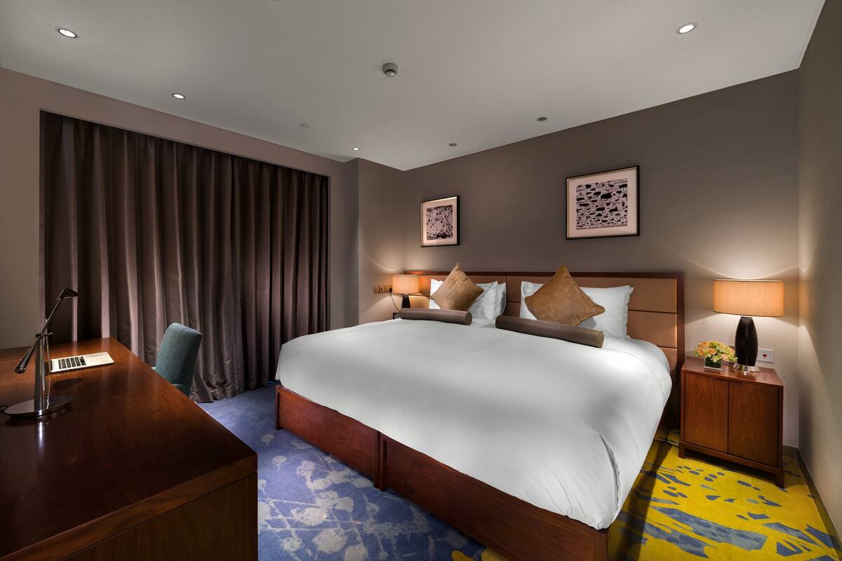 Ein Hotelzimmer mit einem großen Bett, einem Schreibtisch mit Lampe und einem Sessel. An der Wand hängen zwei gerahmte Bilder.