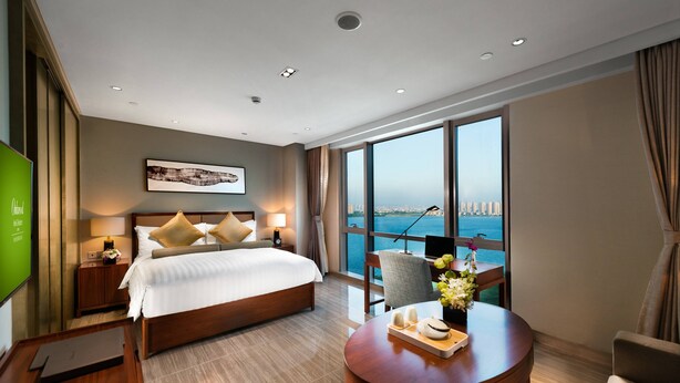 Une chambre d’hôtel moderne dotée d’un grand lit, d’un bureau, d’une chaise et offrant une vue sur la ville et la mer.