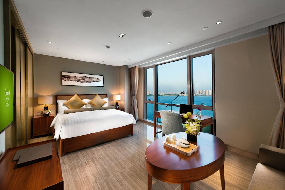 Ein modernes Hotelzimmer mit einem großen Bett, einem Schreibtisch, einem Sessel und Blick auf Stadt und Meer.