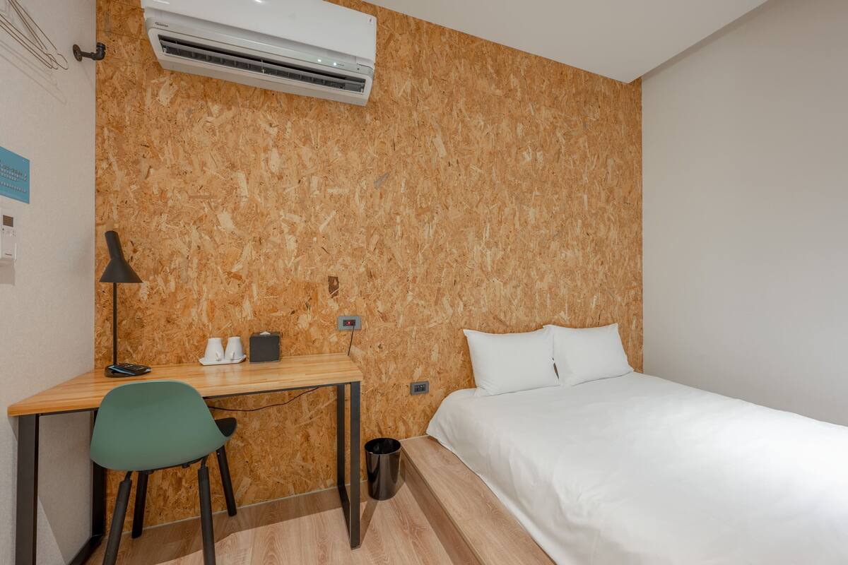 Habitación de hotel con pared de madera, un escritorio con silla, cama con ropa blanca y un equipo de aire acondicionado.