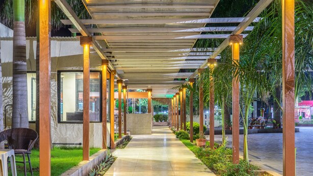 A covered outdoor corridor with a tiled floor, wooden pillars, and a glass wall. - Hotel Pushpak Resort (Rahata)