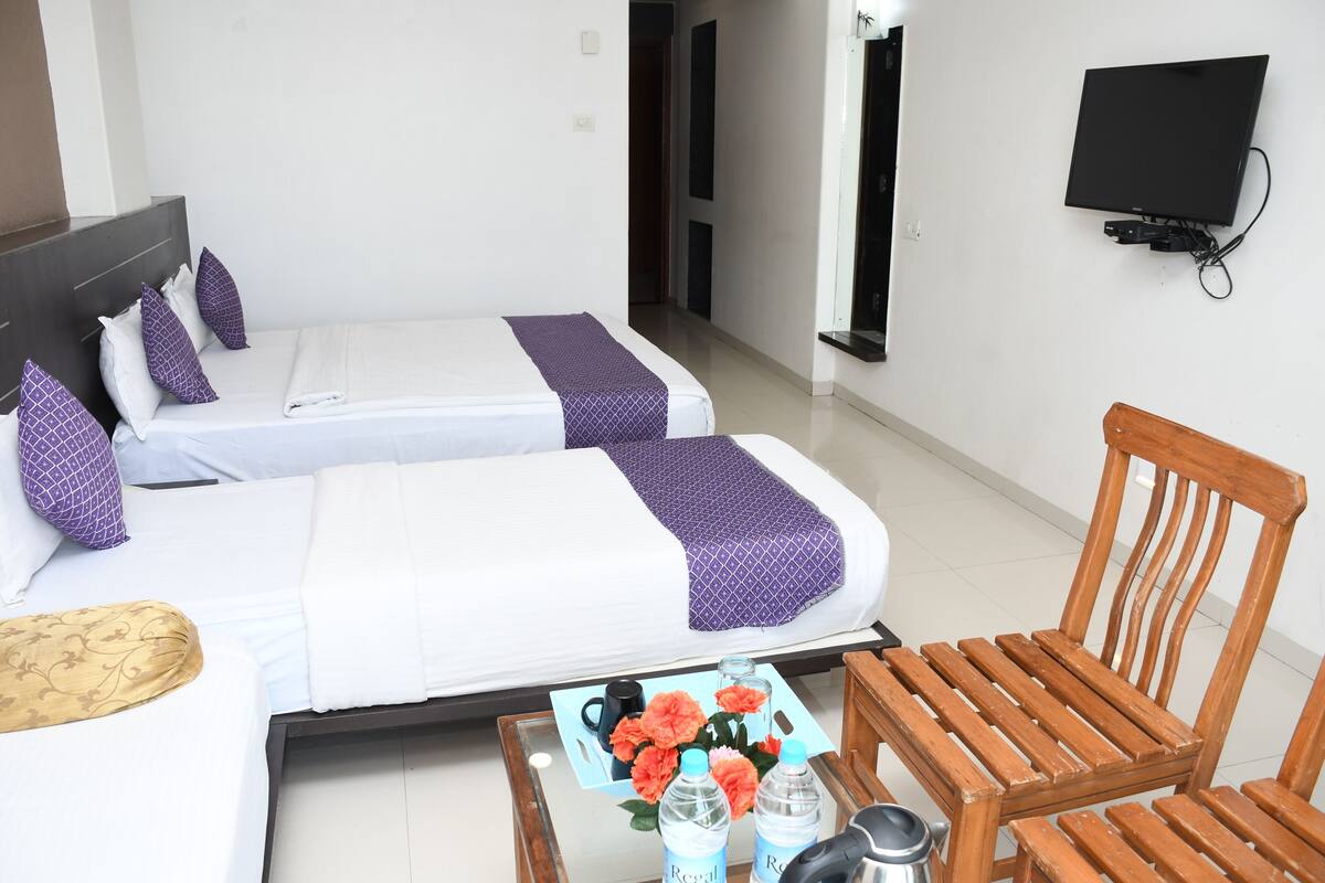 Une chambre d’hôtel avec deux lits, une chaise en bois, une table ornée de fleurs et de bouteilles, et un téléviseur fixé au mur.