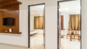 A hotel room with a wooden reception desk, a flat-screen TV, a bed, a bathroom with a shower, and a dining area with chairs and a table. - Hotel Pushpak Resort (Rahata)