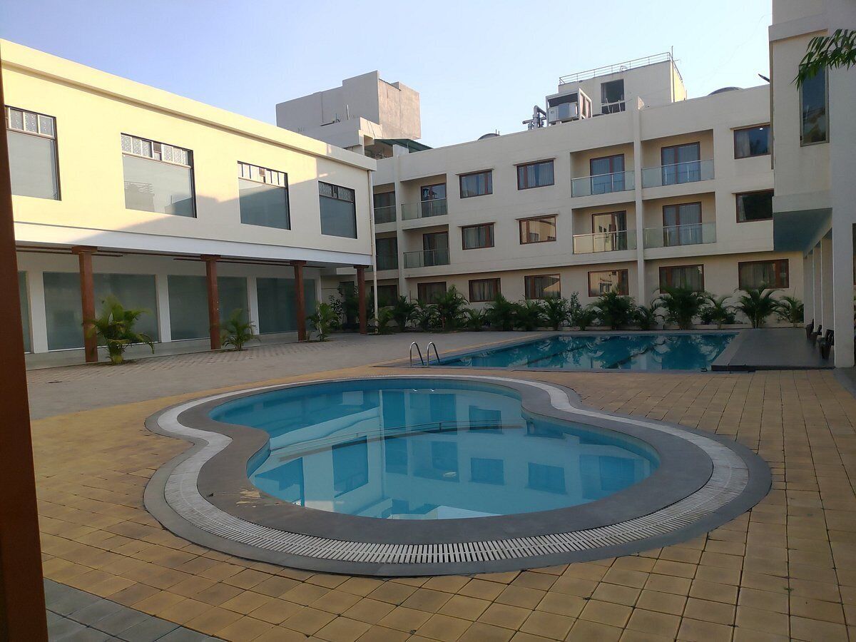 A swimming pool surrounded by a courtyard with tiled flooring and a multi-story building in the background.