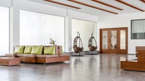 A modern lobby with a sectional sofa, two hanging chairs, a wooden reception desk, and large windows. - Hotel Pushpak Resort (Rahata)