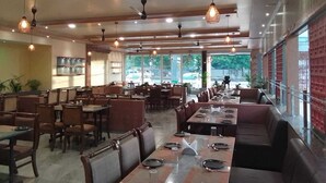 A spacious restaurant with multiple dining areas, including booths and tables, and large windows offering views of the outside. - Hotel Pushpak Resort (Rahata)