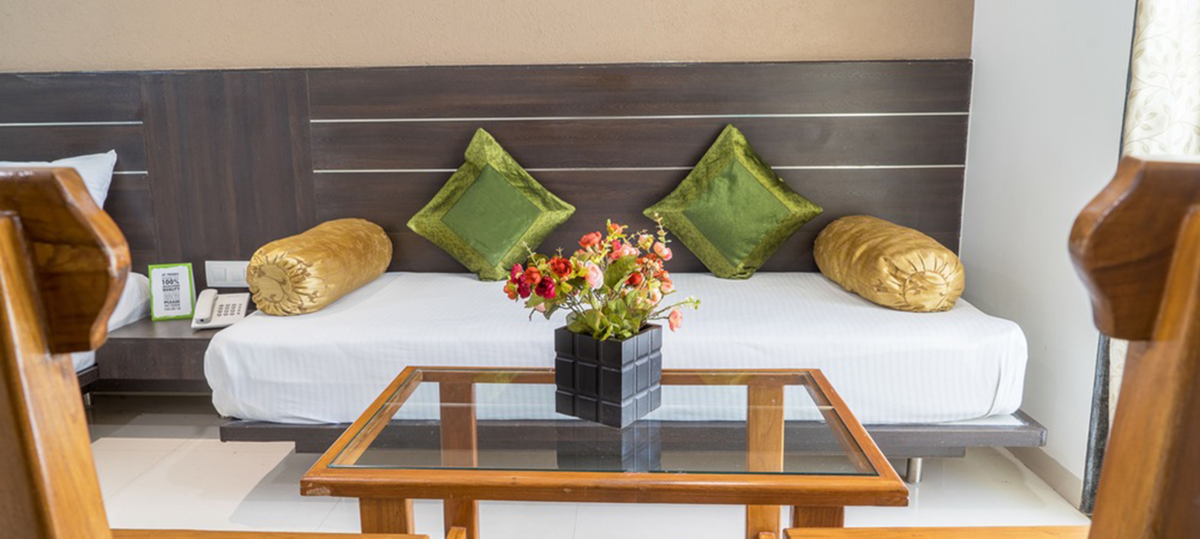 A modern hotel room with a sofa bed, two green pillows, a wooden coffee table with a vase of flowers, and a small bedside table with a lamp.