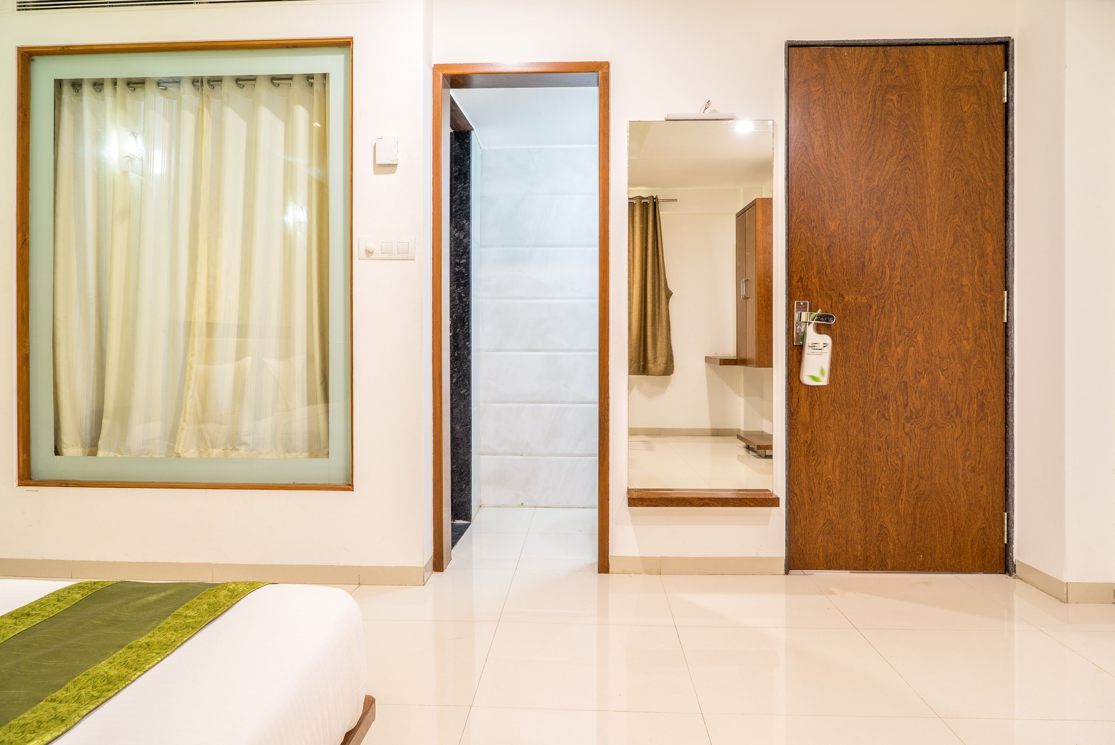 A hotel room with a bed, a large window with curtains, a wooden door, and a bathroom visible through an open door.