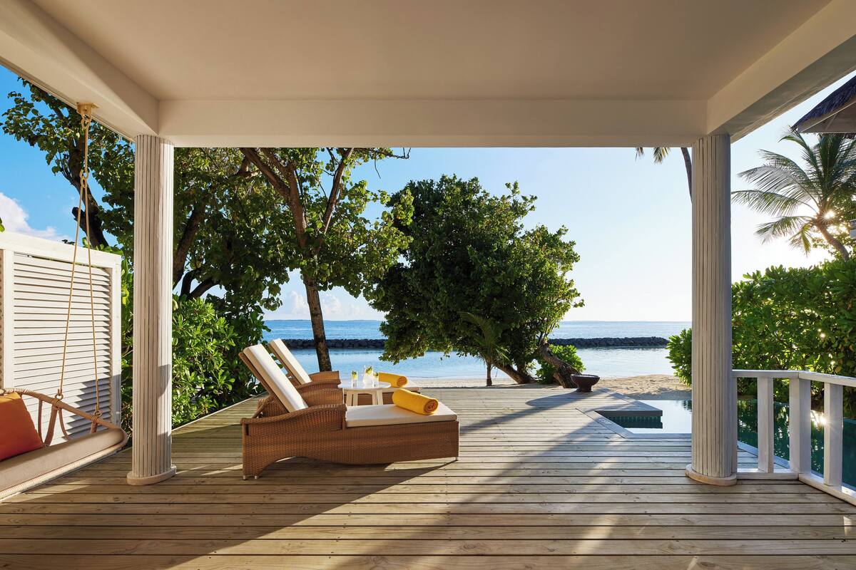 A wooden deck with a wicker lounge chair and a small table, overlooking a beach and ocean.