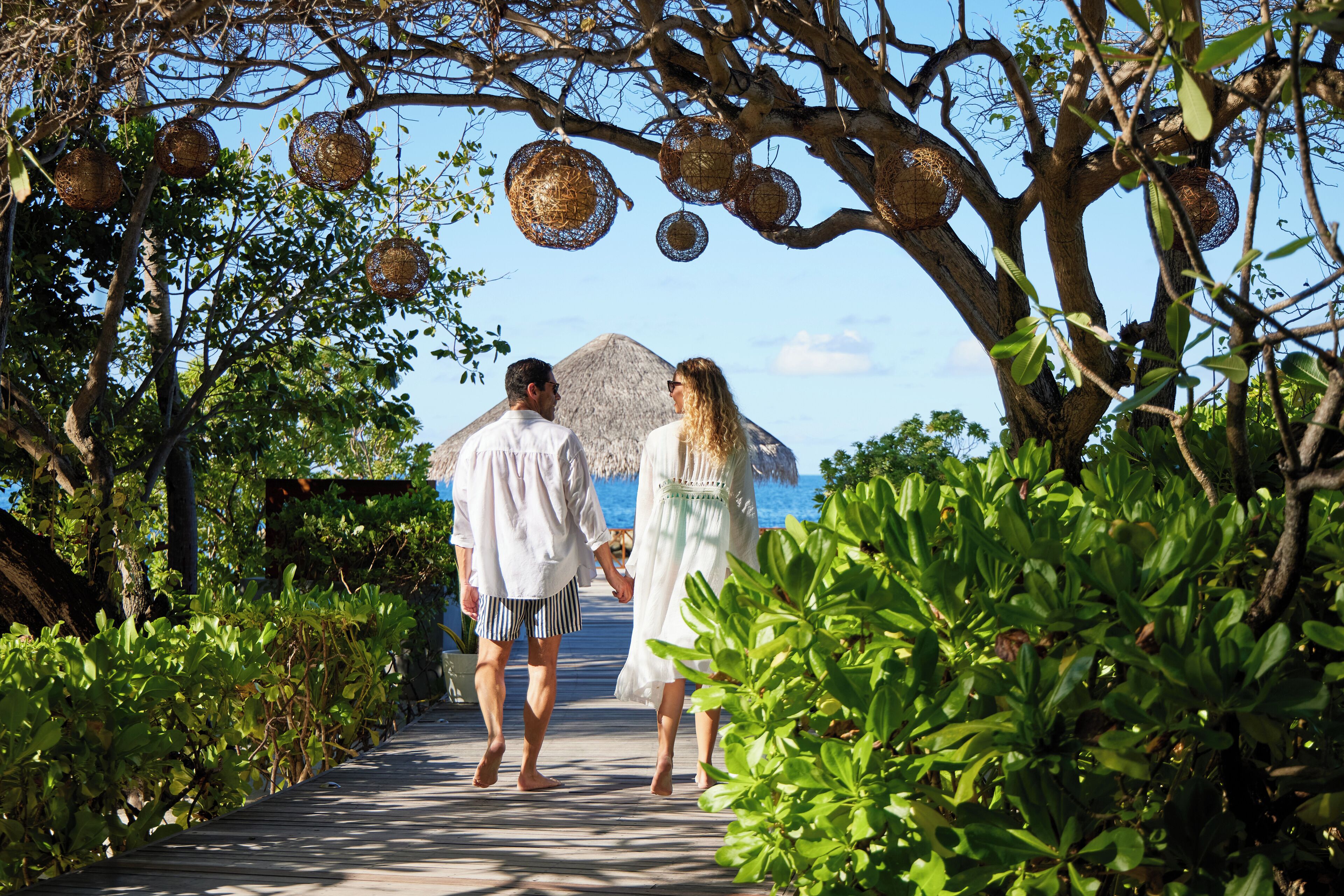 A couple walking on a wooden pathway surrounded by lush greenery and hanging decorative baskets.