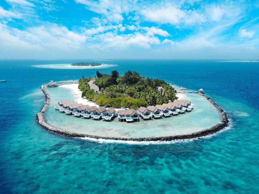 Un complexe hôtelier construit sur pilotis, avec des hébergements de type bungalow, une eau turquoise cristalline et une petite île à la végétation luxuriante.