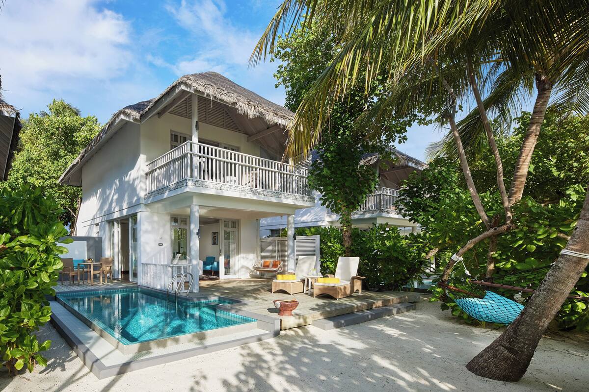 A two-story house with a swimming pool, outdoor seating, and a thatched roof.