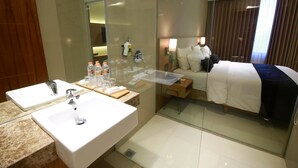Habitación de hotel con un separador de vidrio que divide el baño y el dormitorio. El baño tiene dos lavamanos y un espejo. El dormitorio cuenta con una cama, una mesita de noche y una lámpara.