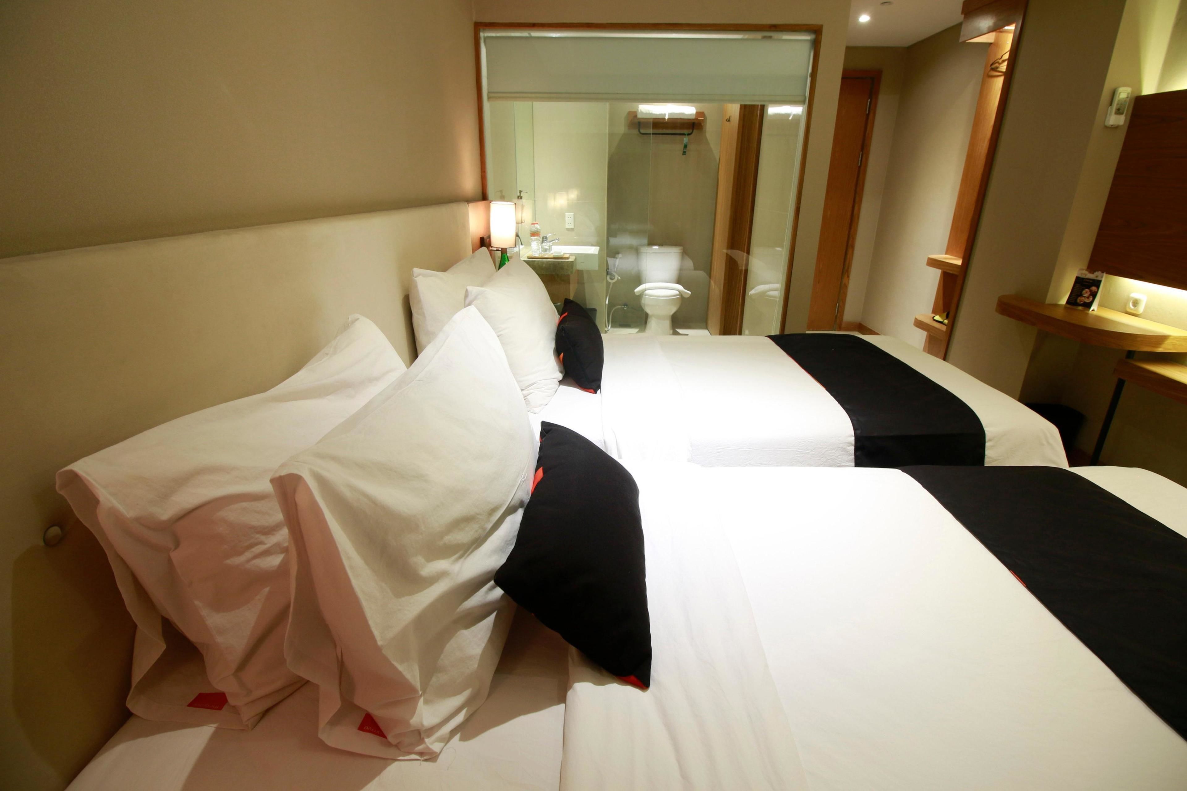 Une chambre d’hôtel avec deux lits, une salle de bain visible à travers une paroi en verre et une petite table avec une lampe.