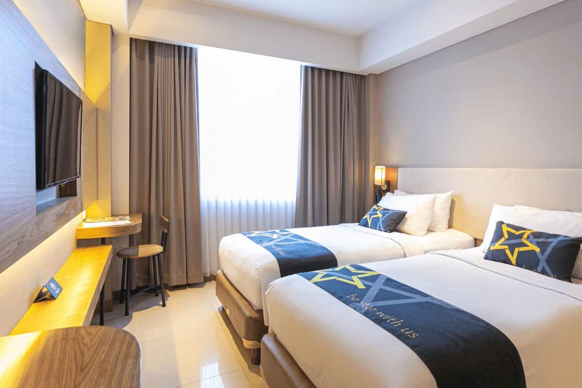 Ein Hotelzimmer mit zwei Betten, einem Schreibtisch, einem Fernseher und einem Fenster mit Vorhängen.