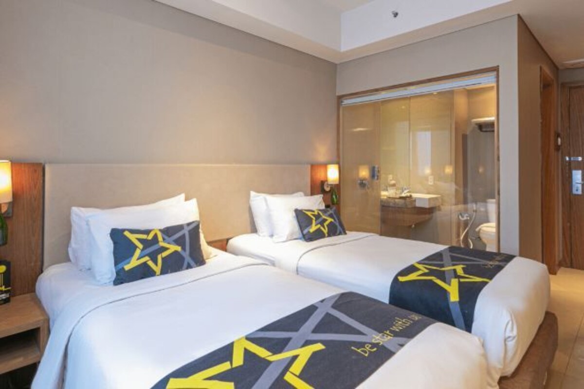 A hotel room with two beds, each with a star-patterned bedspread and matching pillows. There is a glass-enclosed shower area and a wooden door.