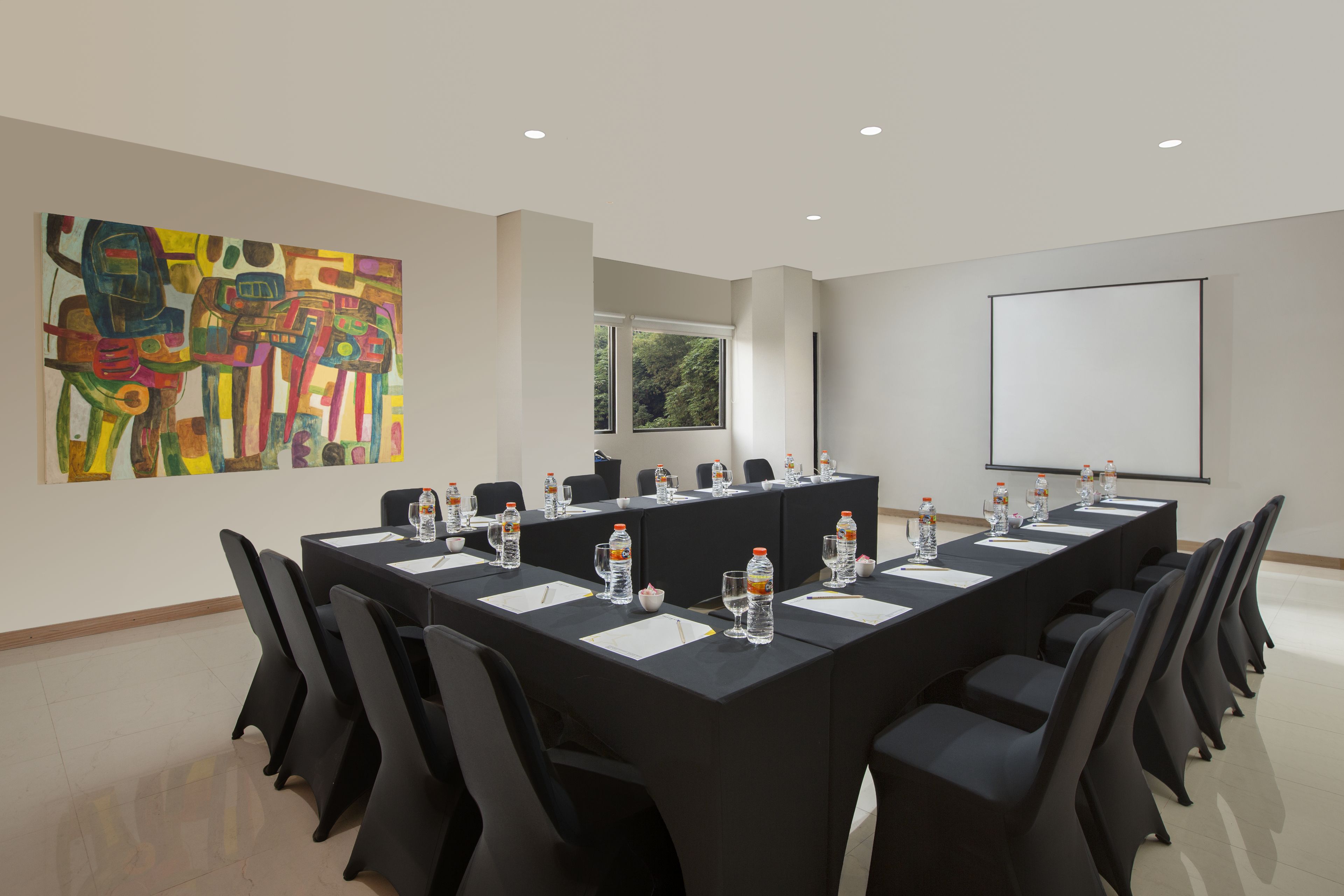 A conference room with a long table, chairs, a projector screen, and a painting on the wall.
