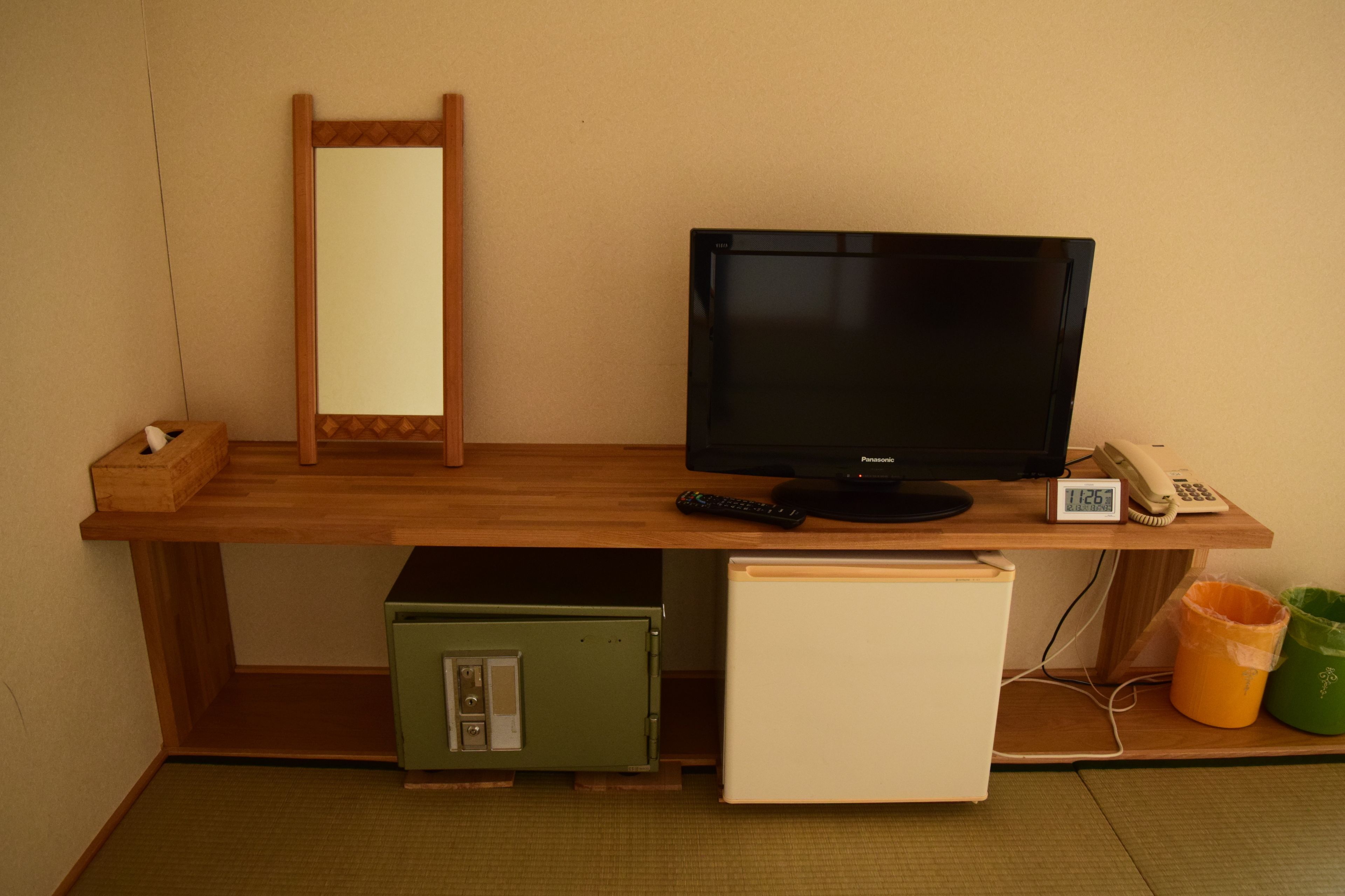 Ein gemütlicher Raum mit Schreibtisch aus Holz, einem schwarzen Fernseher, Telefon und kleinem Kühlschrank.