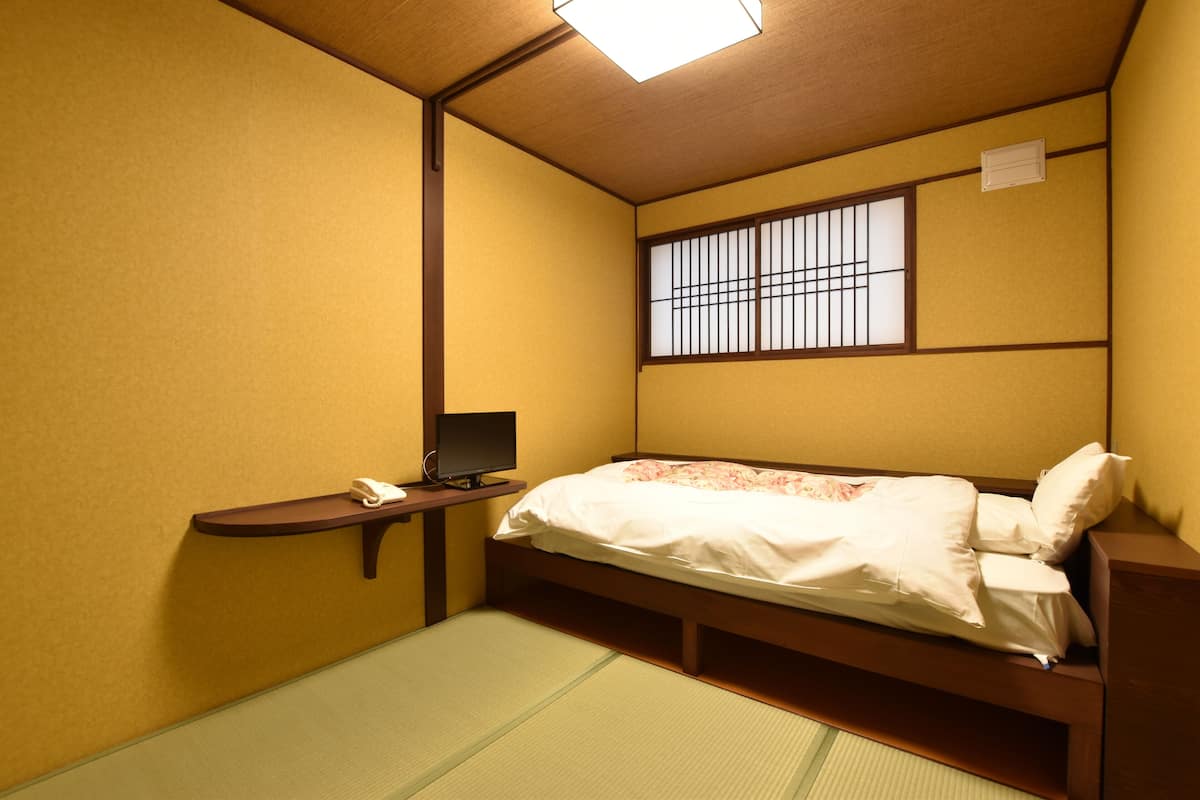 A traditional Japanese-style room with a tatami mat floor, a bed with white bedding, a small desk with a telephone, and a window with bars.