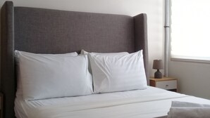 A neatly made bed with white linens and a grey headboard, a bedside table with a lamp, and an air conditioning unit mounted on the wall. - The Concierge at Wind Residences Tagaytay (Tagaytay)