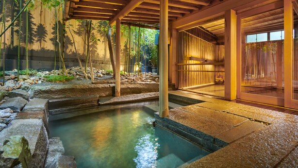 Un espace de bains à l’eau chaude de style japonais traditionnel, avec des marches en pierre menant à un petit bassin, entouré de plantes de bambou et d’une pergola en bois.