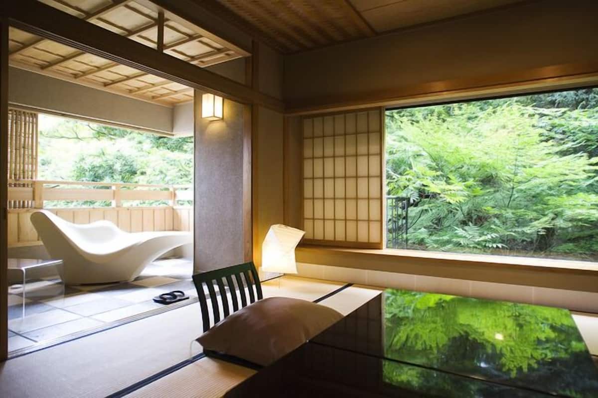 A traditional Japanese room with tatami flooring, a low table, a cushioned chair, and a large window overlooking greenery.