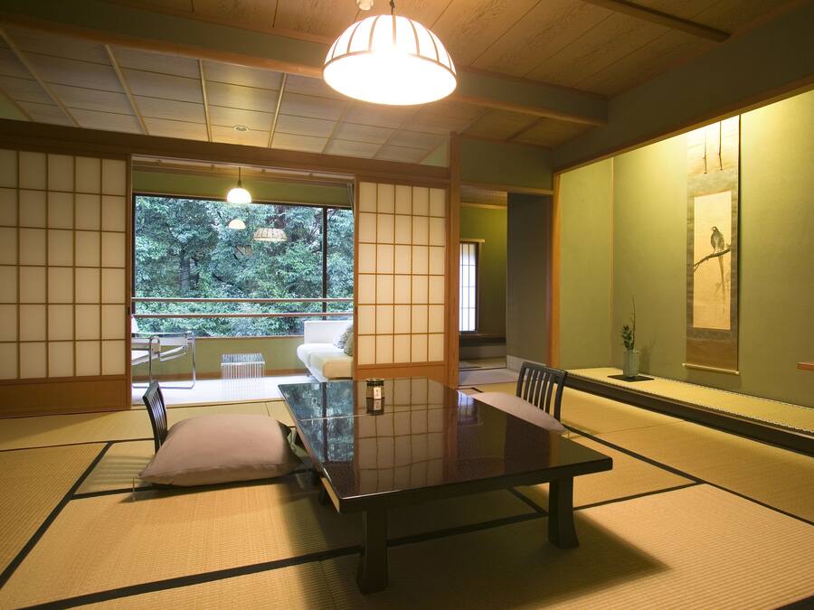 A traditional Japanese room with tatami flooring, a low table, and a sliding door leading to a balcony with a view of trees.