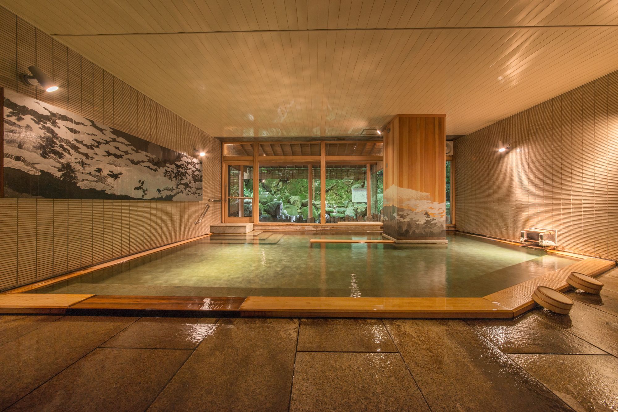 Un onsen japonés tradicional con interior de madera, suelo de piedra y vistas a un paisaje natural a través de puertas correderas.