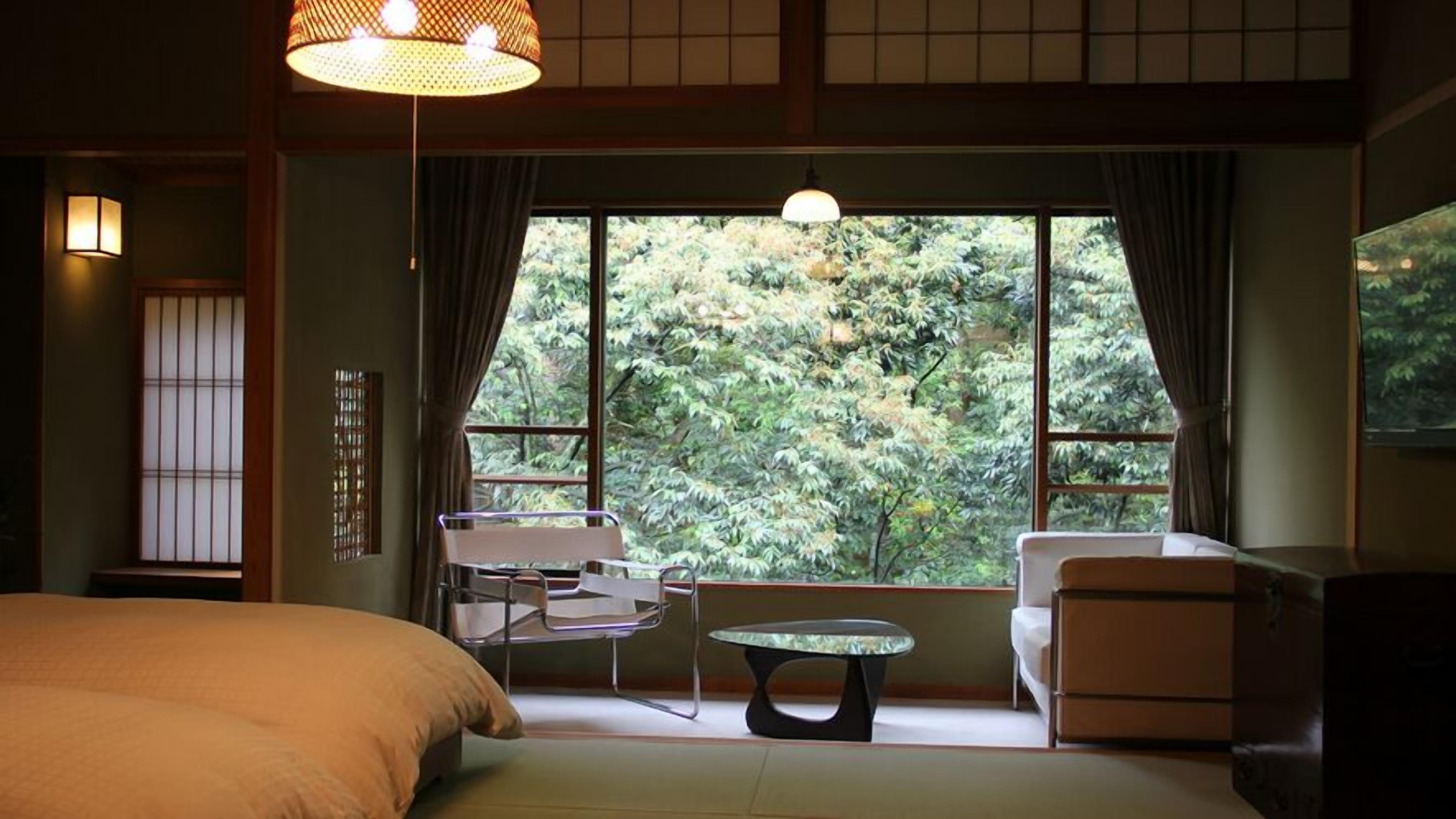 A room with a bed, a table, two chairs, and a view of trees through large windows.