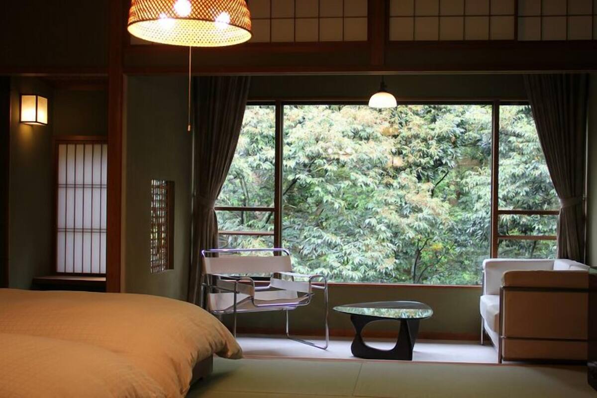A room with a bed, a table, two chairs, and a view of trees through large windows.