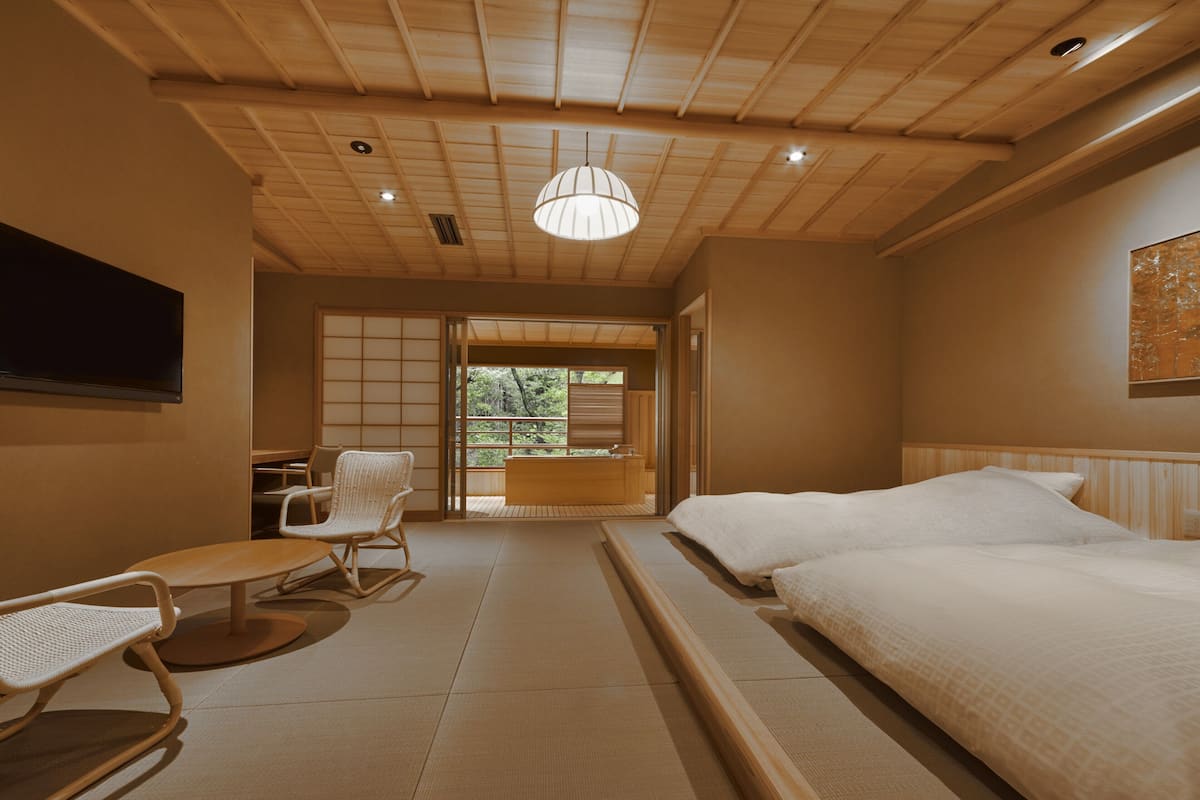 A traditional Japanese-style room with tatami flooring, a low wooden ceiling, and a sliding door leading to a balcony. There are two beds with white bedding and a small table with chairs.